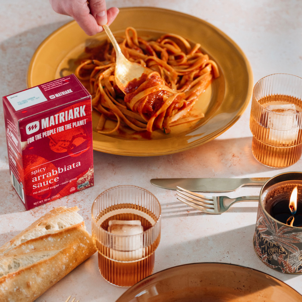 A fork in a plate of pasta, with a carton of Matriark sauce in front of it.